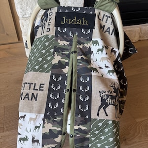 May include: A baby car seat cover with a patchwork design. The cover features various patterns including camouflage, arrows, and deer silhouettes. The name "Judah" is embroidered on a black patch. The cover has a green trim and button closure.