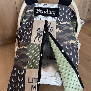 May include: A baby car seat cover with a patchwork design featuring deer, arrows, and camouflage patterns in shades of brown, green, and gray. The cover includes the name "Bradley" on a black label.