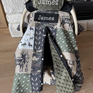 May include: A baby car seat cover with a patchwork design featuring deer, arrows, and camouflage patterns in shades of green, brown, and beige. The cover includes the name "James" embroidered in white on two separate panels.