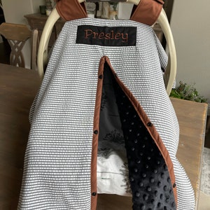 May include: A baby car seat cover with a gray and white striped pattern. The cover has a brown trim and a black interior with a textured, dotted design. The name "Presley" is embroidered in orange on a black patch.