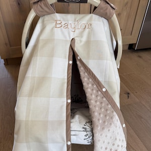 May include: A beige and white plaid baby car seat cover with the name "Baylor" embroidered in brown thread. The cover has a brown trim and a soft, dotted interior. The Graco handle is visible.