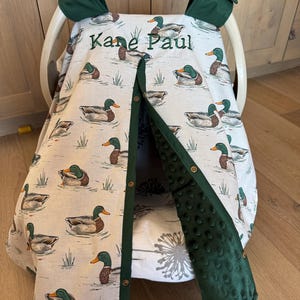 May include: A baby car seat cover with a duck pattern. The cover is cream-colored with illustrations of ducks swimming in water. The trim and accents are dark green. The name "Kane Paul" is embroidered on the cover.