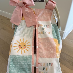 May include: A baby car seat cover with a patchwork design. The cover features a large pink bow with the name "Dallas" embroidered on it. The cover has a "You Are My Sunshine" theme with a smiling sun graphic.