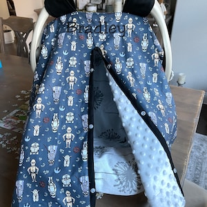 May include: A navy blue baby car seat cover featuring a Star Wars character pattern. The cover has a black trim and a soft, white, textured interior. The name "Bradley" is embroidered on the cover.