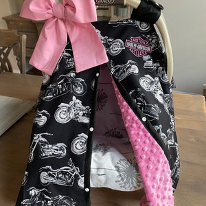 May include: A black baby car seat cover featuring a motorcycle print with a large pink bow. The cover has a pink, textured interior and the Harley-Davidson Cycles logo.