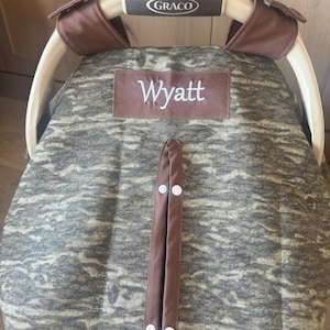 Puede incluir: Una funda para asiento de coche de beb&eacute; con un estampado de camuflaje en tonos verdes, marrones y grises. La funda tiene un ribete marr&oacute;n y un parche bordado con el nombre "Wyatt". El asa del asiento del coche tiene la marca "Graco".