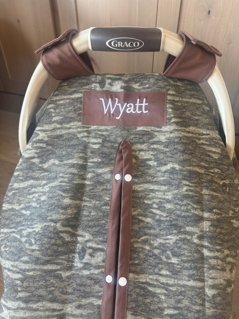 Puede incluir: Una funda para asiento de coche de beb&eacute; con un estampado de camuflaje en tonos verdes, marrones y grises. La funda tiene un ribete marr&oacute;n y un parche bordado con el nombre "Wyatt". El asa del asiento del coche tiene la marca "Graco".