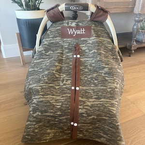 Puede incluir: Una funda para asiento de coche de beb&eacute; con un estampado de camuflaje en tonos marrones y grises. La funda tiene un parche de cuero marr&oacute;n con el nombre "Wyatt" bordado en blanco. Correas marrones con broches blancos aseguran la funda.