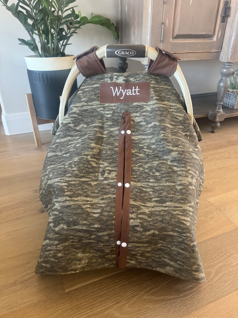Puede incluir: Una funda para asiento de coche de beb&eacute; con un estampado de camuflaje en tonos marrones y grises. La funda tiene un parche de cuero marr&oacute;n con el nombre "Wyatt" bordado en blanco. Correas marrones con broches blancos aseguran la funda.