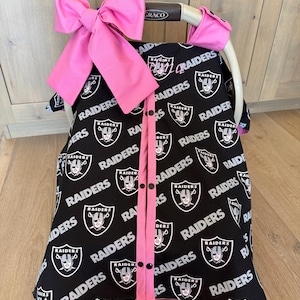 May include: A black baby car seat cover featuring the Raiders logo in white. The cover has a pink bow and trim. The cover is designed to fit a Graco car seat.