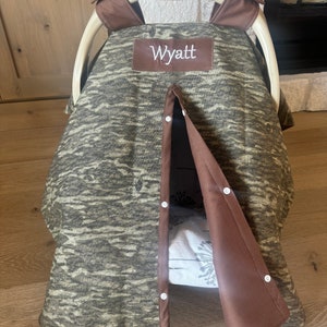 Puede incluir: Una funda para asiento de coche de beb&eacute; con un estampado de camuflaje en tonos verdes y marrones. La funda tiene un ribete marr&oacute;n y el nombre "Wyatt" bordado en un parche marr&oacute;n. La funda est&aacute; parcialmente abierta, revelando un interior estampado.