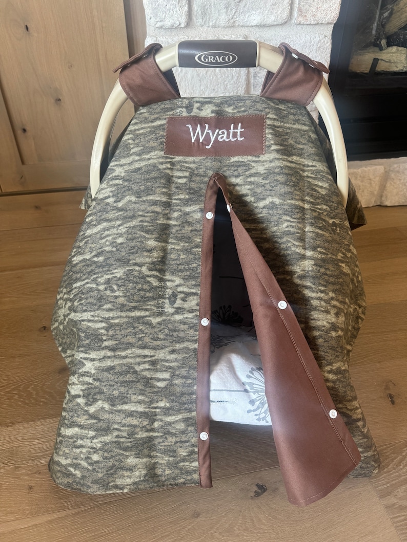 Puede incluir: Una funda para asiento de coche de beb&eacute; con un estampado de camuflaje en tonos verdes y marrones. La funda tiene un ribete marr&oacute;n y el nombre "Wyatt" bordado en un parche marr&oacute;n. La funda est&aacute; parcialmente abierta, revelando un interior estampado.