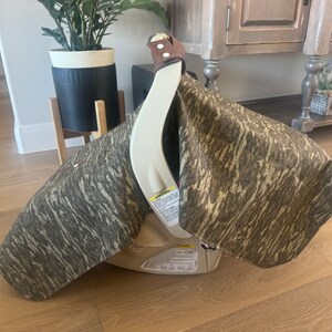 Puede incluir: Asiento de coche beige con una funda con estampado de camuflaje. La funda est&aacute; hecha de una tela con tonos marrones, grises y beige. El asiento del coche tiene una correa de cuero marr&oacute;n y est&aacute; sobre un suelo de madera.