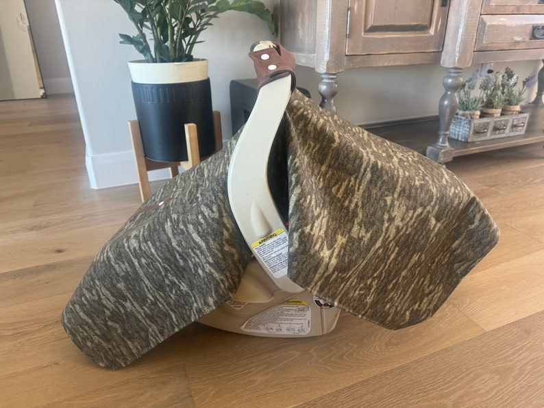 Puede incluir: Asiento de coche beige con una funda con estampado de camuflaje. La funda est&aacute; hecha de una tela con tonos marrones, grises y beige. El asiento del coche tiene una correa de cuero marr&oacute;n y est&aacute; sobre un suelo de madera.