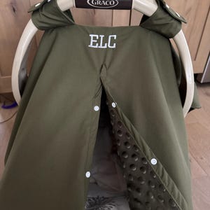May include: An olive green baby car seat cover with the letters "ELC" embroidered in white. The cover has a button closure and a soft, textured interior. The car seat handle is brown with the brand name "Graco".