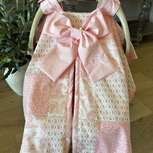 May include: A pink and white patterned car seat cover featuring a large central bow with the embroidered name Jaxlyn. The fabric displays a patchwork design with floral motifs and western-themed illustrations, secured with white snaps on the front.