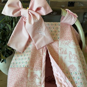 May include: A pink baby car seat cover featuring a patchwork design with floral patterns and western-themed horse illustrations. A large, matching pink fabric bow is attached to the top, embroidered with the name Joxlyn in white script lettering.