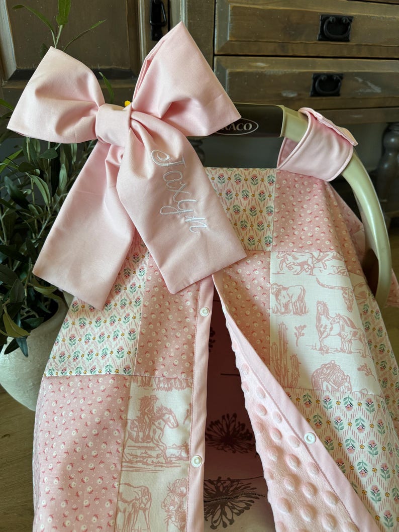 May include: A pink baby car seat cover featuring a patchwork design with floral patterns and western-themed horse illustrations. A large, matching pink fabric bow is attached to the top, embroidered with the name Joxlyn in white script lettering.