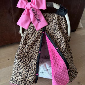 May include: A baby car seat cover with a leopard print design. The cover has a pink bow with the name "Belaia" embroidered on it. The interior is pink and the edges are black.