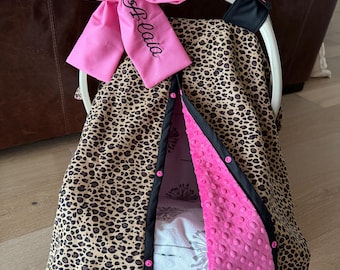 May include: A baby car seat cover with a leopard print design. The cover has a pink bow with the name "Belaia" embroidered on it. The interior is pink and the edges are black.