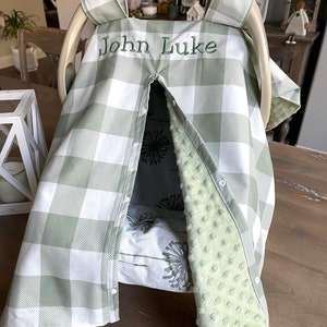 May include: A baby car seat cover with a green and white checkered pattern. The cover has the embroidered name "John Luke" in green thread. The interior features a floral print and a soft, green, textured fabric.