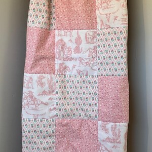 May include: A patchwork quilt featuring alternating squares of pink dimpled fabric, a white fabric with a pink western-themed print of cowboys, horses, and cacti, and a white fabric with a small repeating floral pattern in pink, green, and blue.