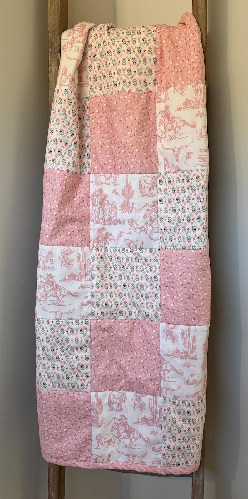 May include: A patchwork quilt featuring alternating squares of pink dimpled fabric, a white fabric with a pink western-themed print of cowboys, horses, and cacti, and a white fabric with a small repeating floral pattern in pink, green, and blue.