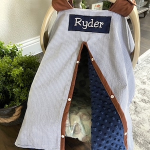 May include: A blue and white striped car seat cover with the name "Ryder" embroidered on a navy blue patch. The cover has brown trim and a navy blue, textured interior. The car seat is a Graco brand.