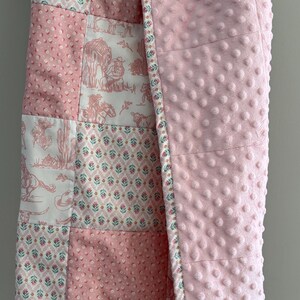 May include: A patchwork baby blanket featuring a mix of pink and white fabrics with floral patterns and western-themed illustrations of cowboys, horses, and cacti. The reverse side is a solid light pink fabric with a raised dimpled texture.