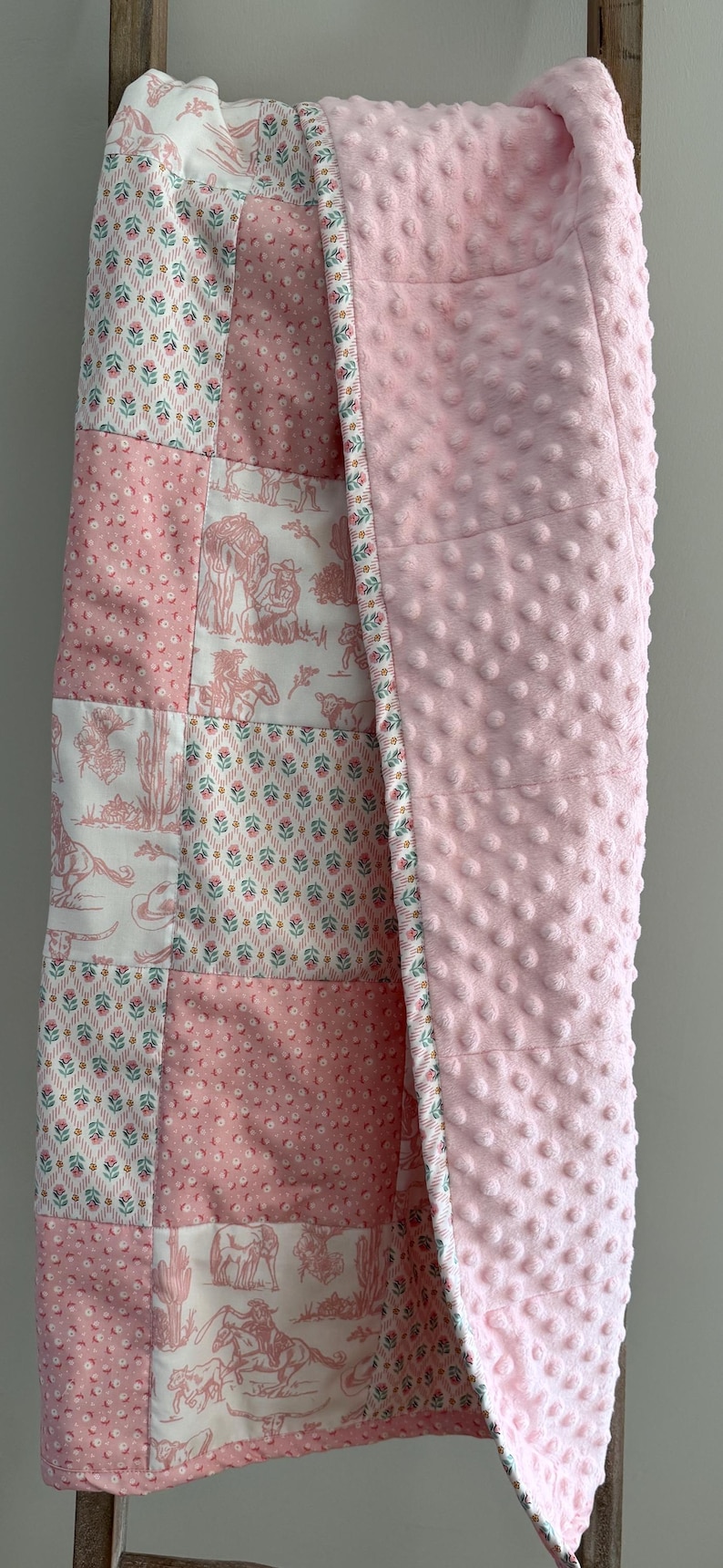 May include: A patchwork baby blanket featuring a mix of pink and white fabrics with floral patterns and western-themed illustrations of cowboys, horses, and cacti. The reverse side is a solid light pink fabric with a raised dimpled texture.