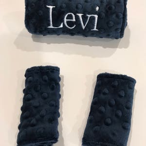 May include: Navy blue, textured fabric baby car seat strap covers. The set includes a long, rectangular cover embroidered with the name "Levi" in white, and two smaller strap covers. The fabric has a soft, plush feel.