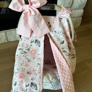 May include: A baby car seat cover with a floral pattern in pink, gray, and white. The cover has a large pink bow with the name "Isabella" embroidered on it. The cover is designed to fit a Graco car seat.