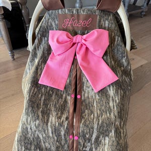 May include: A baby car seat cover with a camouflage pattern, a large pink bow, and the name "Hazel" embroidered on a brown patch. The cover has brown trim and pink buttons.