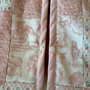 May include: A patchwork garment featuring a mix of fabrics in shades of pink and white. The design includes panels with floral patterns, geometric shapes, and western-themed illustrations of horses and cacti. Two white plastic snap buttons are visible.