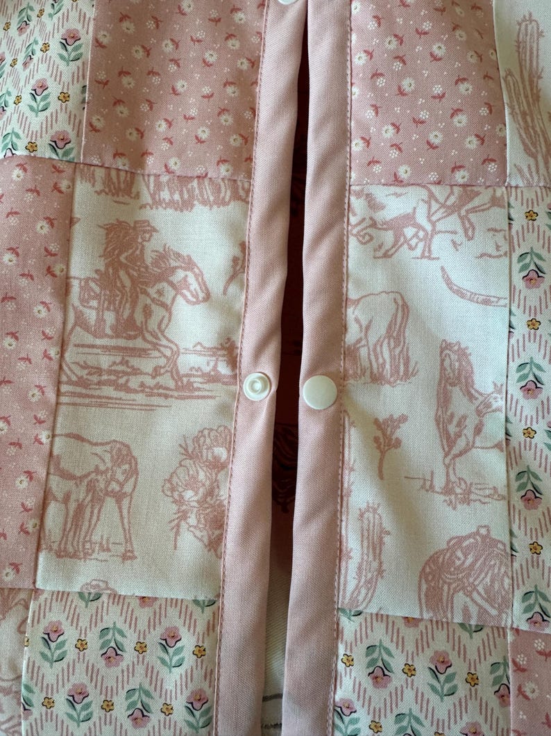 May include: A patchwork garment featuring a mix of fabrics in shades of pink and white. The design includes panels with floral patterns, geometric shapes, and western-themed illustrations of horses and cacti. Two white plastic snap buttons are visible.