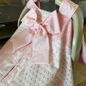 May include: A pink and white patterned baby car seat cover draped over a Graco carrier. The fabric features a mix of floral, horse, and cow skull prints with textured pink minky panels. A large pink bow with the embroidered name Joxyn is attached.