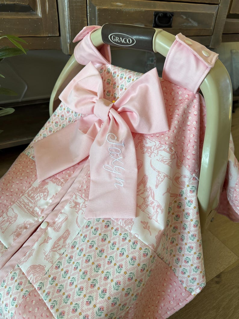 May include: A pink and white patterned baby car seat cover draped over a Graco carrier. The fabric features a mix of floral, horse, and cow skull prints with textured pink minky panels. A large pink bow with the embroidered name Joxyn is attached.