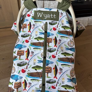 May include: A custom baby car seat cover with a fishing theme. The white fabric is patterned with fish, fishing rods, and boats. The name "Wyatt" is embroidered on the top. The cover has a green trim and is attached to a Graco car seat.
