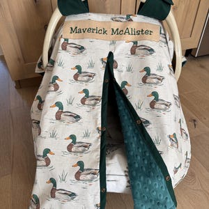 May include: A baby car seat cover with a duck pattern. The cover is cream-colored with illustrations of ducks swimming in water. The trim and interior lining are a dark green. The name "Maverick McAlister" is embroidered on the cover.
