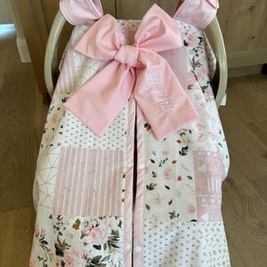 May include: A pink and white baby car seat cover with a large pink bow. The cover features a patchwork design with floral and geometric patterns. The name "Annie" is embroidered on the bow.