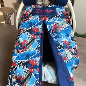 May include: A baby car seat cover featuring a Spider-Man design. The cover is primarily blue with a repeating pattern of Spider-Man figures and cityscapes. The name "Xavier" is embroidered in red. The cover has a navy blue trim and red snap closures.