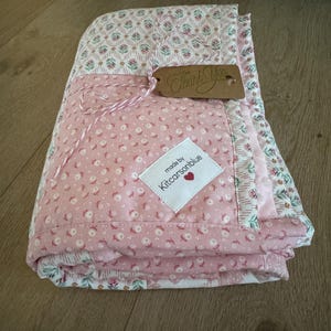 May include: A folded quilt featuring a pink fabric with a small white floral pattern and a white fabric with a pink and green floral print. A white label reads made by Kitcarsonblue with a red heart, and a brown tag says Thank You.