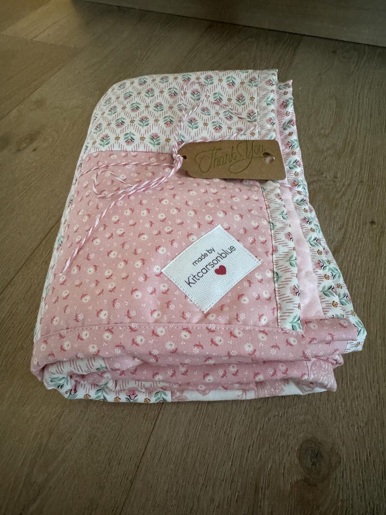 May include: A folded quilt featuring a pink fabric with a small white floral pattern and a white fabric with a pink and green floral print. A white label reads made by Kitcarsonblue with a red heart, and a brown tag says Thank You.