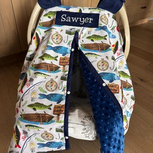 May include: A baby car seat cover with a fishing theme. The white fabric is patterned with fish, boats, fishing poles, and compasses. The name "Sawyer" is embroidered on a navy blue patch. The cover has a navy blue trim and a soft, blue, textured lining.