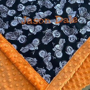May include: A black and orange baby blanket with a motorcycle pattern. The blanket features a black background with white motorcycle illustrations. The name "Jason Dale" is embroidered in orange thread. The border is orange with a textured, dotted pattern.