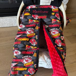 May include: A baby car seat cover featuring a black background with a repeating pattern of red and yellow Kansas City Chiefs logos and text. The interior is red, and the cover is personalized with the name "Matthew".