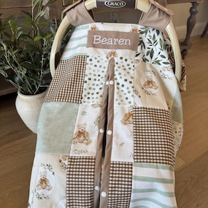 May include: A baby car seat cover with a patchwork design. The cover features squares of brown gingham, green, and white fabric with teddy bear and floral patterns. The word "Bearen" is embroidered on a tan patch.