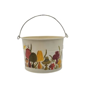 May include: A white metal bucket with a silver handle. The bucket is decorated with a painted design of colorful mushrooms.