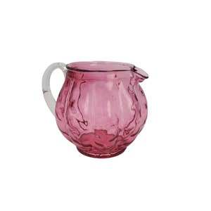 May include: A pink glass pitcher with a clear glass handle. The pitcher has a textured, bubbled surface.
