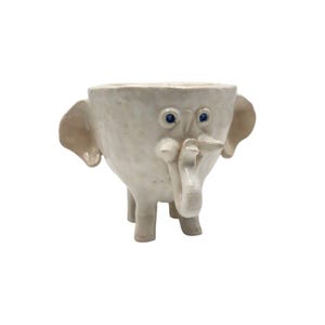 May include: A quirky, cream-coloured ceramic elephant planter with blue eyes. The planter features a trunk, ears, and four legs. The elephant-shaped planter is designed to hold small plants or flowers, adding a touch of playfulness to any space.